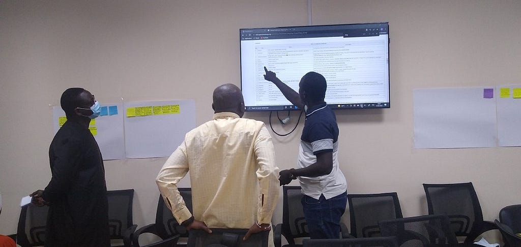 Three Senegalese participants discussing data validation and naming conventions in front of a big screen TV and with flip chart paper on the wall.