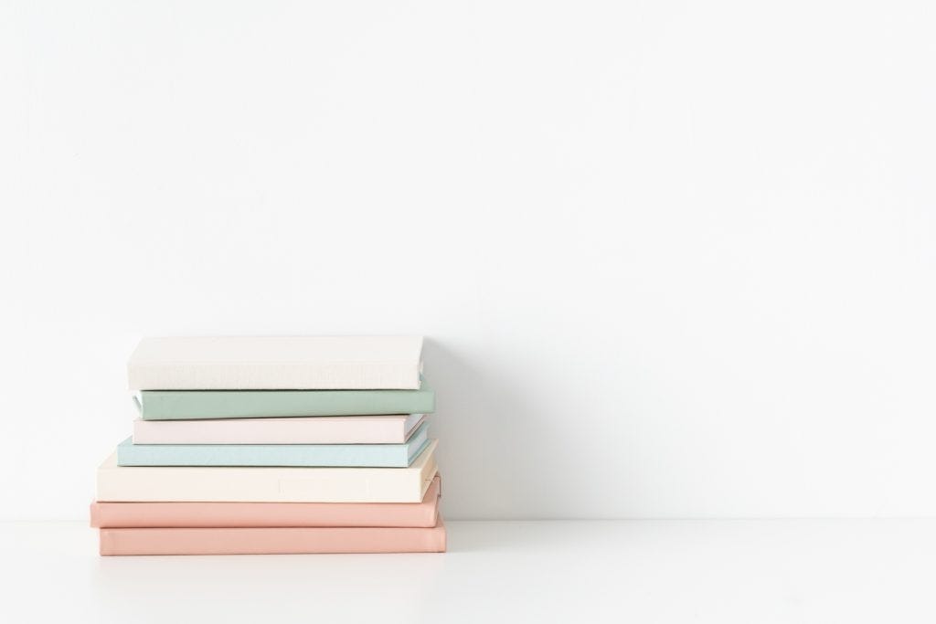 A stack of pastel-colored books arranged neatly against a white background. The books are in shades of pink, green, and beige