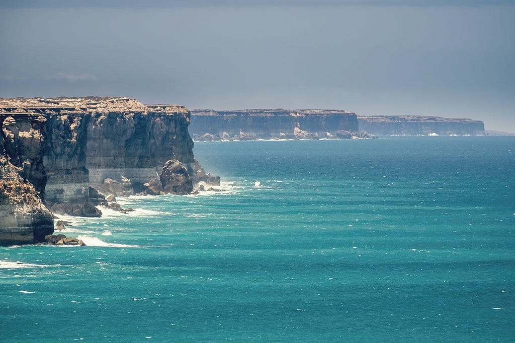 View of the Great Australian Bight, featuring the expansive blue ocean with waves crashing against the rugged cliffs under a hazy sky.