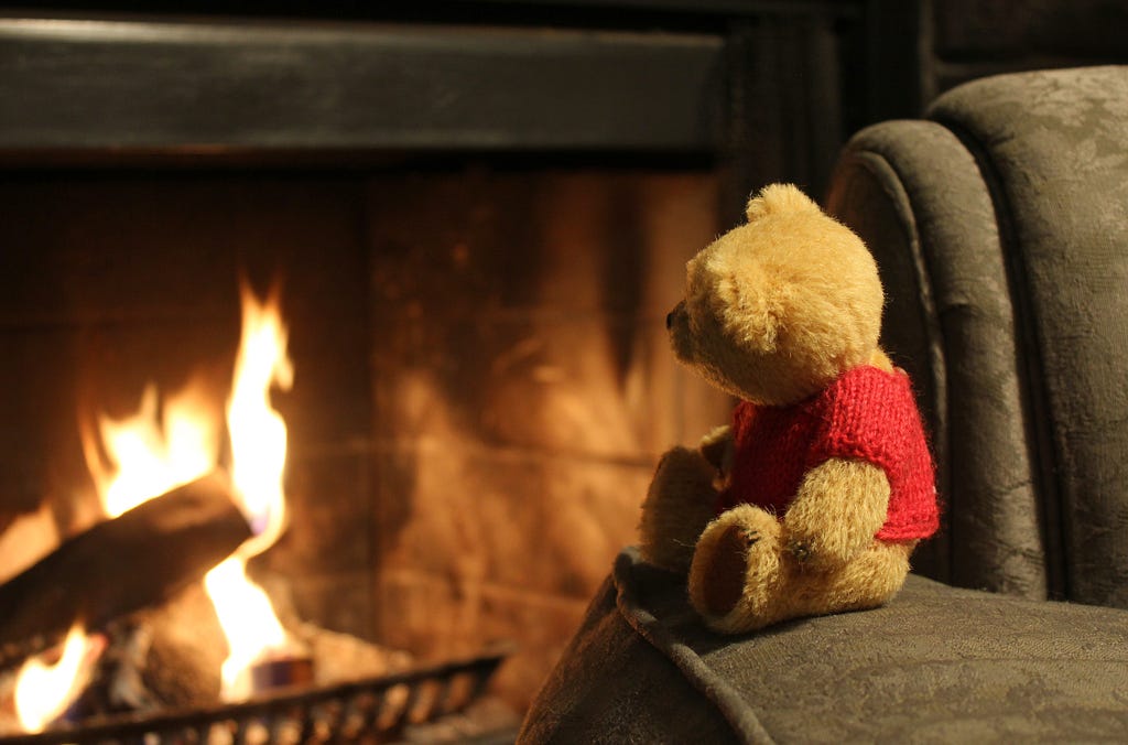 A teddy bear in a red sweater sitting on a sofa looking at a nearby fireplace