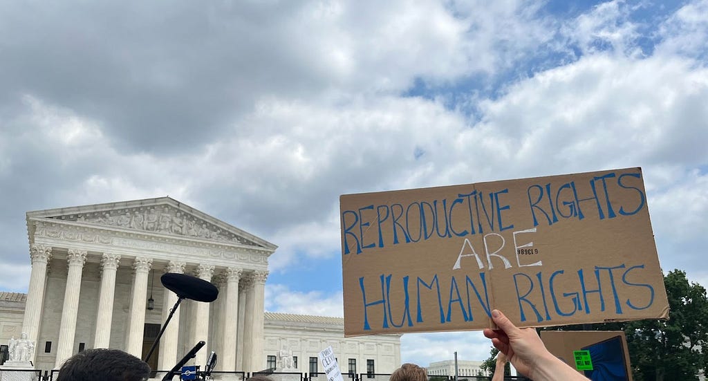 An image of a protest happening in front of a courthouse. One hand is raising a sign that says ‘Reproductive rights are human rights’. In the background, we can see the sky and dark clouds.