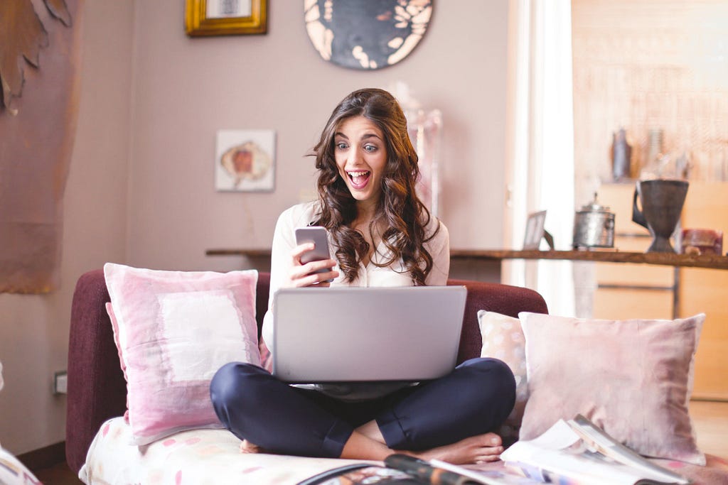 Woman sitting with laptop in her lap and excitedly looking at her phone