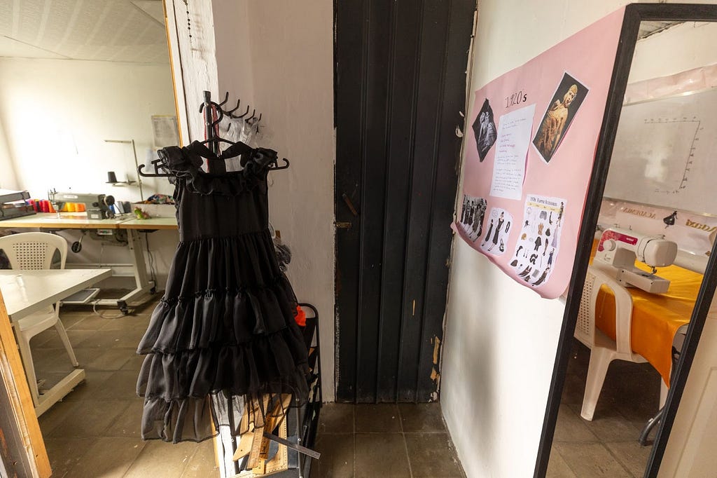 A sleeveless black dress with ruffles hangs on a rack with a sewing machine and work tables in the background.