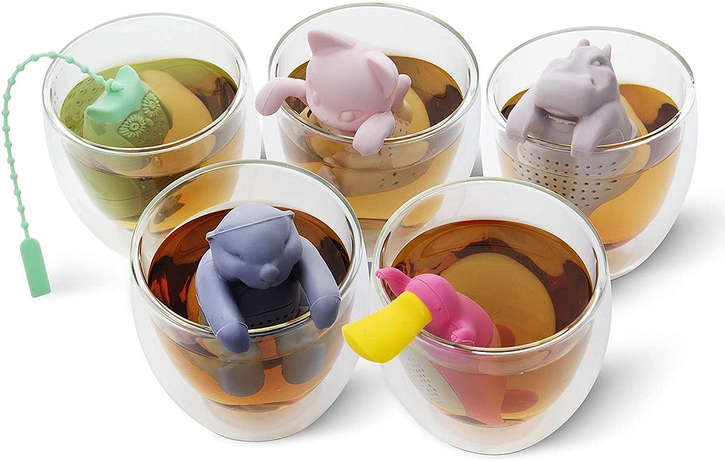 Five glass cups of tea sit on a white countertop. In each cup sits a silicone tea infuser, all in various colors and different animal shapes. In the topmost row, from left to right, is a teal owl, a pink cat, and a dusky purple hippo. In the row below is a blue bear and a pink platypus with a yellow bill.