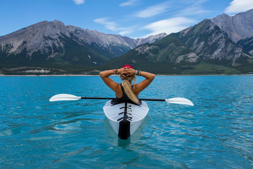 A picture of a lady from the back, seated on a boat and relaxing with her hands interlaced behind her head, parked still in beautiful blue waters and overlooking breathtaking hilly horizon; depicting a break you can catch and the end.