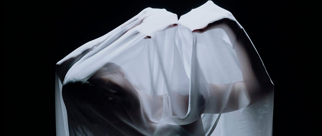 A woman in silhouette draped beneath a long, white, shear veil strikes a pose with hands tented above her tilted head.
