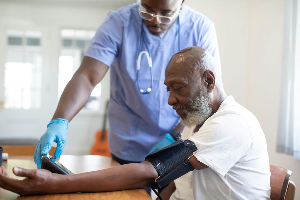 A black man with a grey beard is having his blood pressure measured by a nurse, a black man with glasses who stands behind him. They are both looking at the machine that is providing the reading.