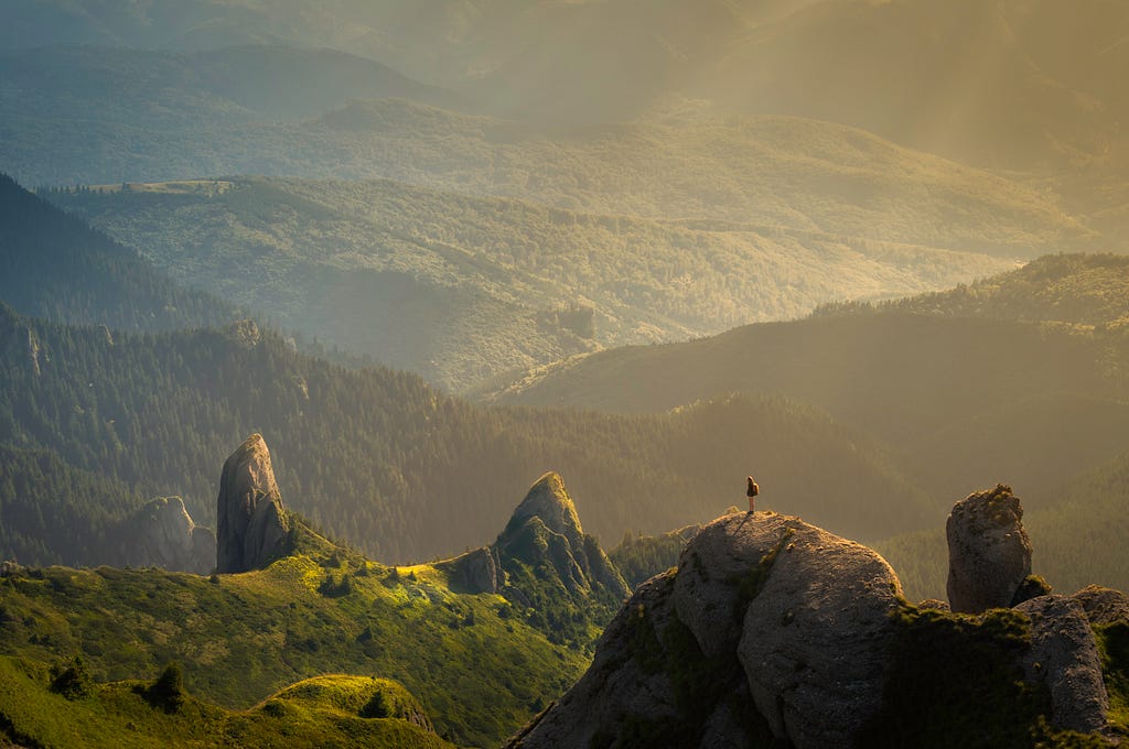 Singular man standing by the edge of a cliff, overlooking rolling hills of lush greenery with sunlight casting its rays upon them.