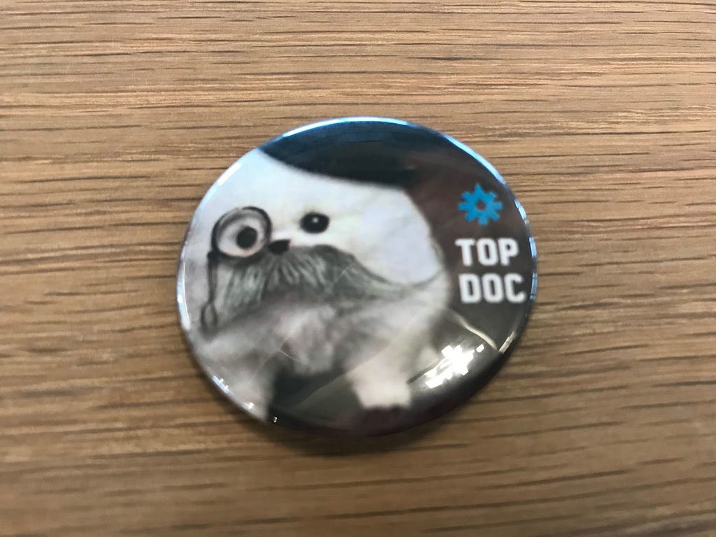 Picture of a button pin, with an image of a small dog with a mustache and a monocle, reading “Top Doc”