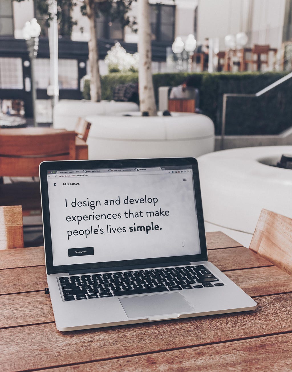 A laptop on a wooden desk and the screen is showing a black text on a white background, saying ‘I design and develop experiences that make people’s lives simple’.