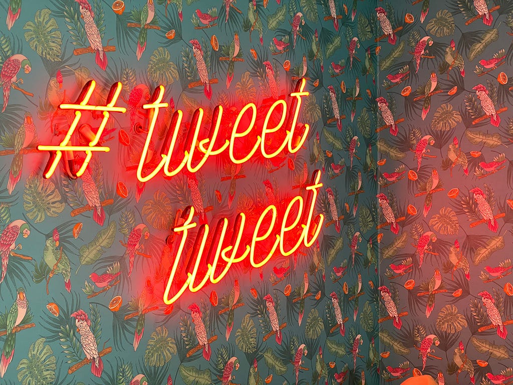 A wall with parrot wallpaper and a neon sign that says “#tweet tweet”
