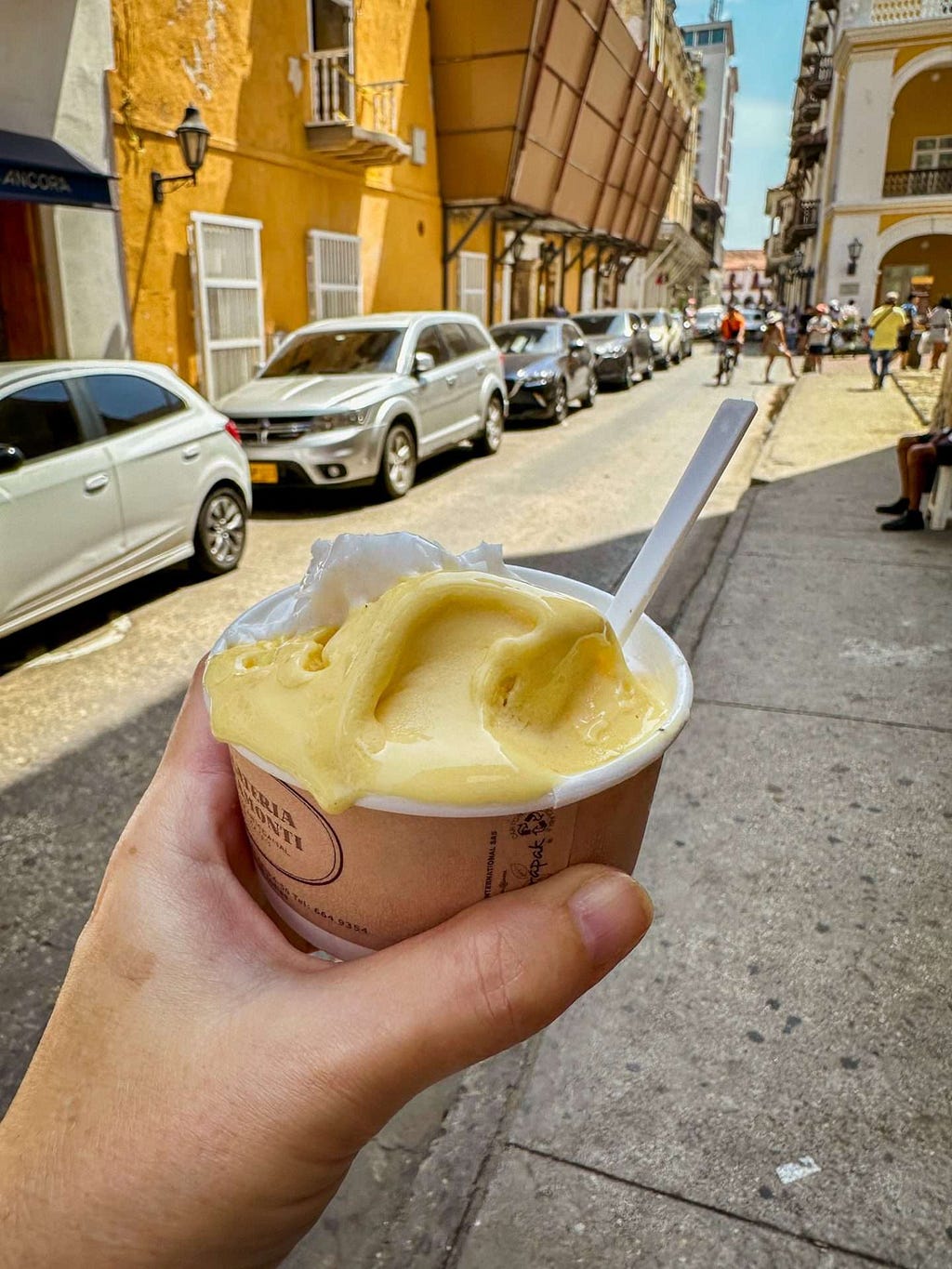 A hand holding a cup of passionfruit and coconut gelato with a white plastic spoon on a sunlit street in Cartagena, Colombia lined with parked cars and buildings.