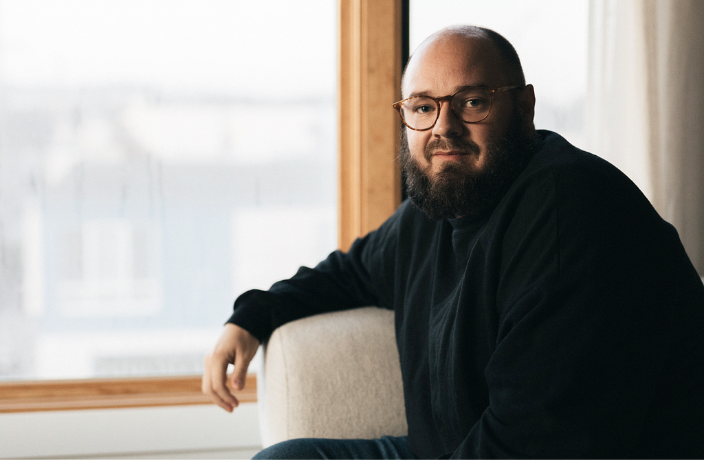 A portrait photo of Tim. He is off-center, looking at the camera. He has a shaved head and a beard, and is wearing a black sweater and glasses. Behind him, there is a window with a wooden frame.