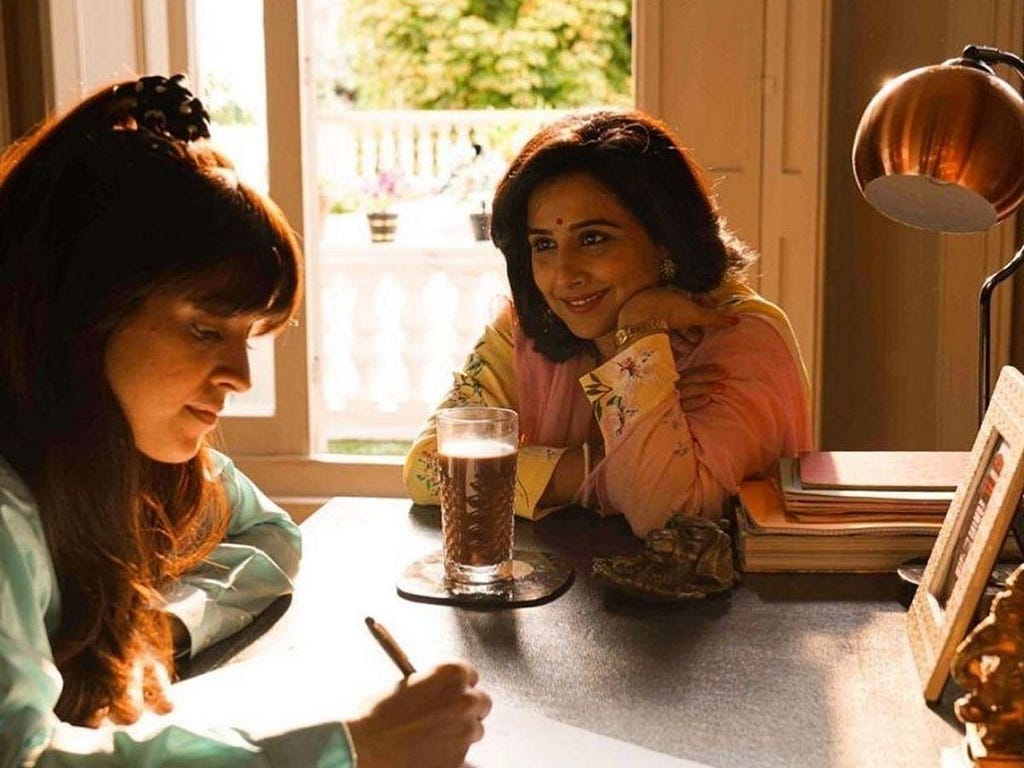 Vidya Balan dressed in a yellow-pink kurti as Shakuntala Devi sits smiling next to Sanya Malhotra who is writing something. There is a glass of water between them.