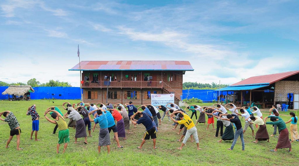 A group stretches arms towards the left in rows, a morning exercise in a rehabilitation program. They stand in the center of a field with the rehabilitation center behind them.