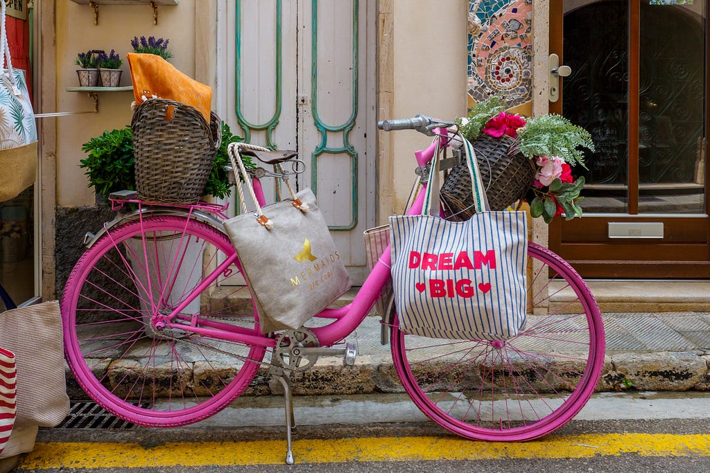 A pink bicycle having plant saplings in front and rear basket. It has bags with “Dream big” written on one of it.