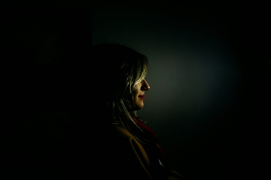 The profile of a woman in the dark. She has a knowing smile on her face as she looks into the light. She is not as dumb as people think she is.