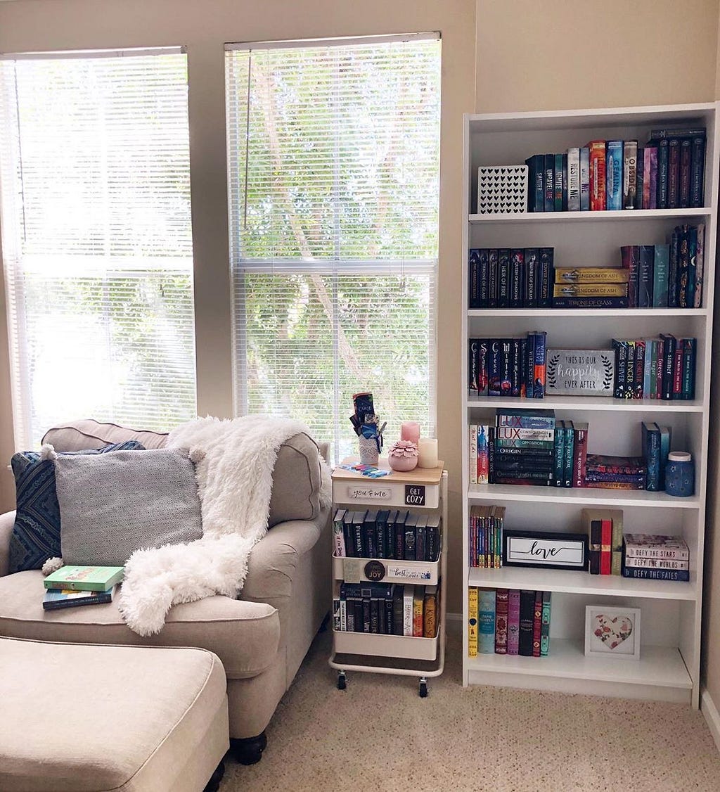 A cozy reading corner with a beige accent chair and ottoman next to a tall bookshelf and rolling cart filled with books.