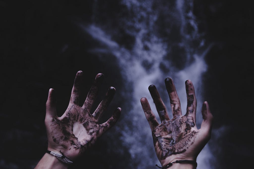 Photo of two hands in front of the camera, palms facing the camera with fingers splayed open, thumbs out and pinky fingers close together. The palms and fingers are dirty with a black-brown substance. Each wrist wears a string bracelet. The background is dark and blurry, with a tumultuous waterfall in the center of the image, well behind the hands.