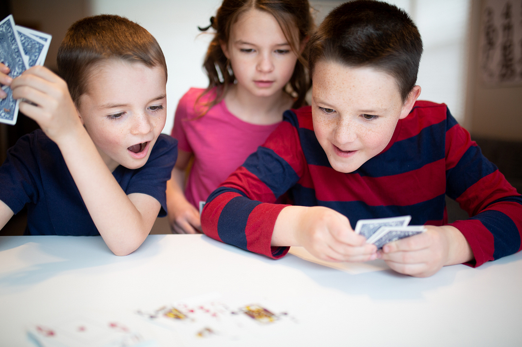 Three children on holidays excitedly playing a card game holding cards and looking down at some spread on the table.