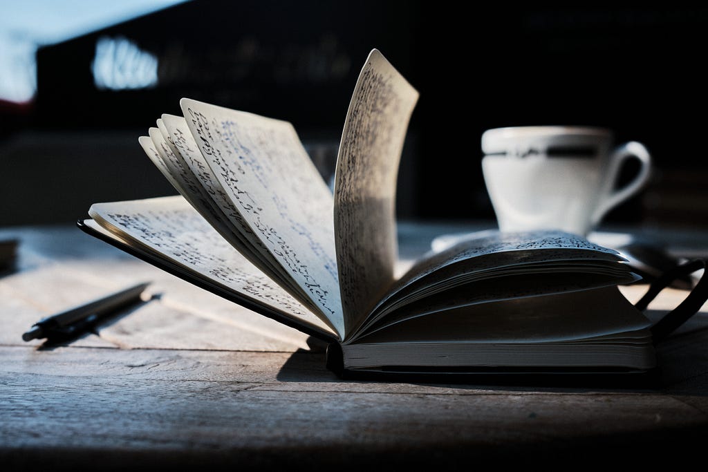 A book sitting on a wooden desk next to a pen and mug. The pages of book are filled with writing and are turning by themselves.