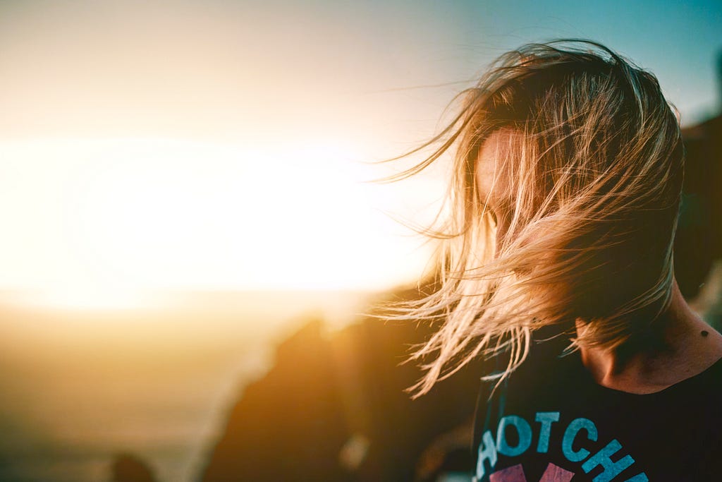 a woman looking down, her hair blown by the wind, in background the sun is setting.