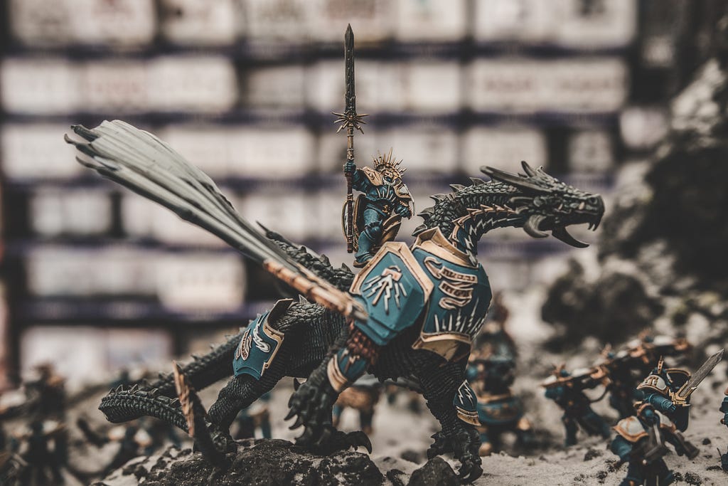 Moody image with shallow depth of field featuring a dragon “action figure” wearing armor and readying for battle. There’s a warrior astride its back and warrior figures surrounding it.