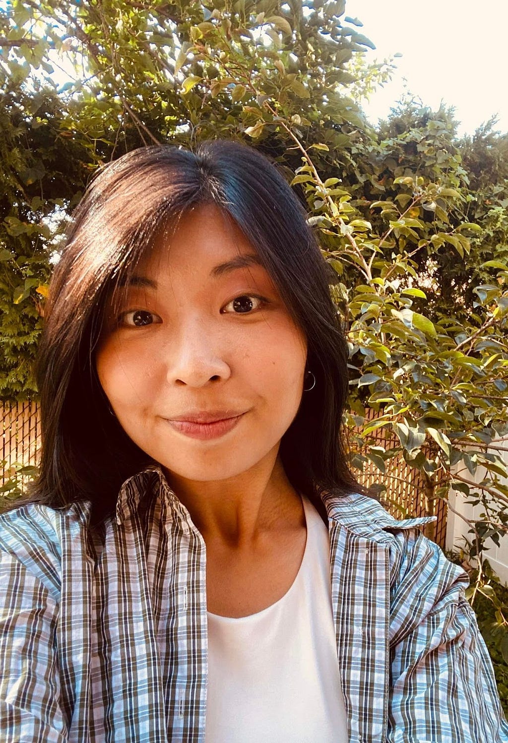 a headshot of Rachel Lim, wearing a blue plaid and white shirt with long black hair. She is smiling in front of a background of sunny fauna and flora.