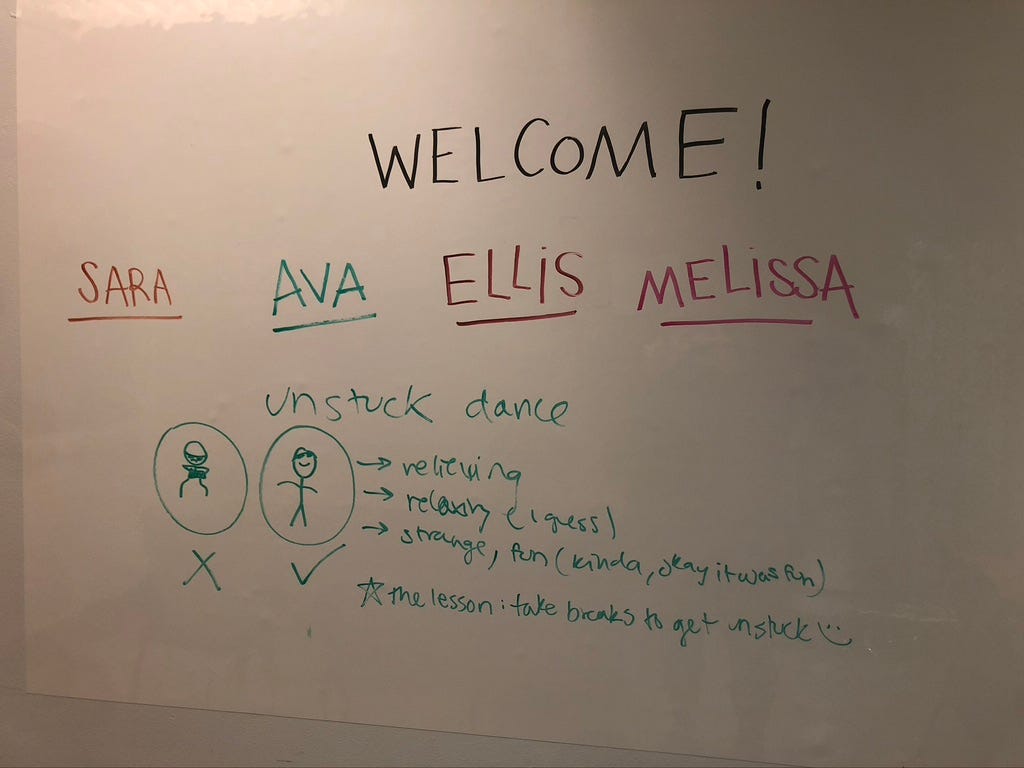 A white board with hand-written words on it. The words read, “Welcome! Sara, Ava, Ellis, Mellisa.” Below this welcome message is a drawing of stick-figures dancing to relieve stress.