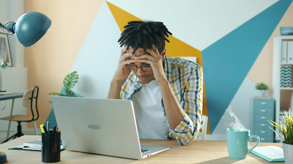 A person sits at a desk holding their head in frustration while looking at a laptop screen. The workspace includes a teal mug with steam rising from it, pens, and a desk lamp. The background wall features bold geometric shapes in teal, mustard yellow, and white, adding a modern feel to the scene. The image conveys stress or difficulty concentrating while working. Photo by Vitaly Gariev on Unsplash
