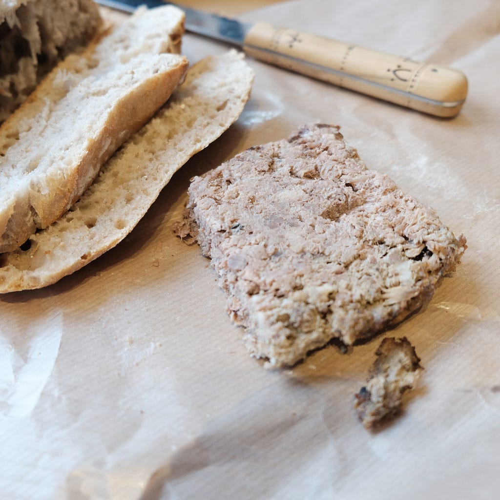 A block of pâté is seen beside a partly sliced loaf of rustic bread. There is also a knife, ready to spread the pâté on the bread— alt text to boost Medium stories