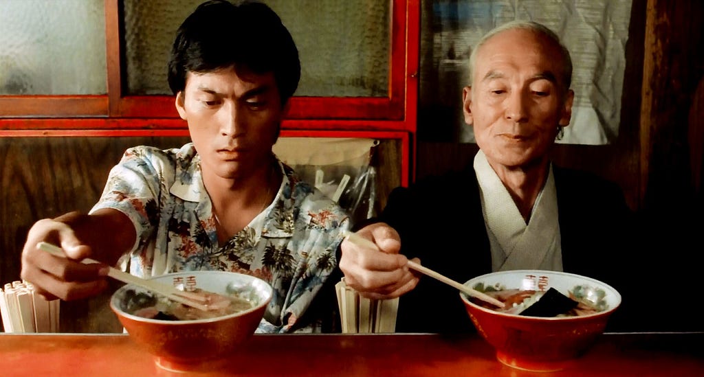 A young and old man sit facing us, staring at their respective bowls of ramen while holding out their chopsticks.