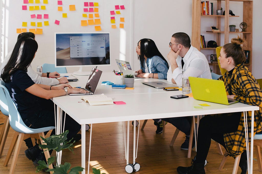 A team of ux designers gather around a table to collectively view a large monitor, which stands in front of a whiteboard of sticky notes.