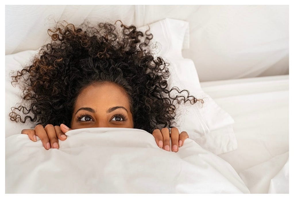 Curly haired woman peeking out from underneath a white duvet