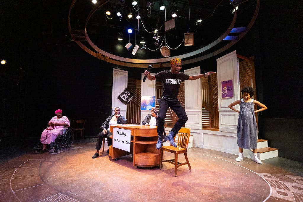 A man in a black t-shirt with the word SECURITY on it jumps off a chair while holding a flashlight and performing. His family members watch from the background.