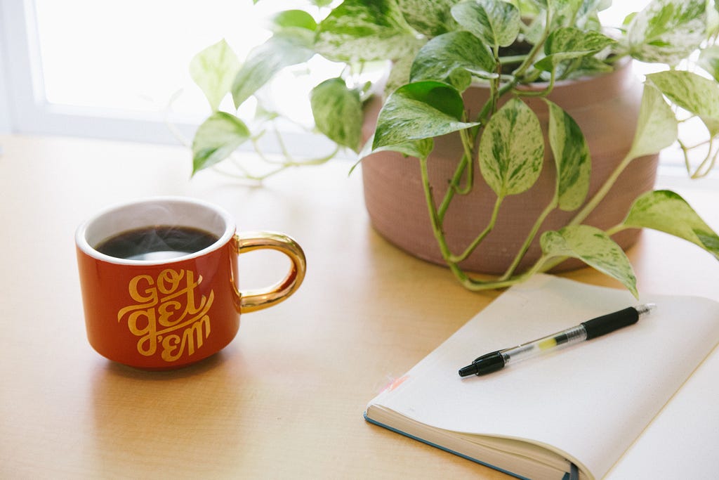 A pothos plant in a ceramic pot, a red coffee mug with a golden handle and text that says, “Go get ‘em,” and an open journal with a black pen on a blank page all lay on a tan, wooden desk in front of a sunlit window.