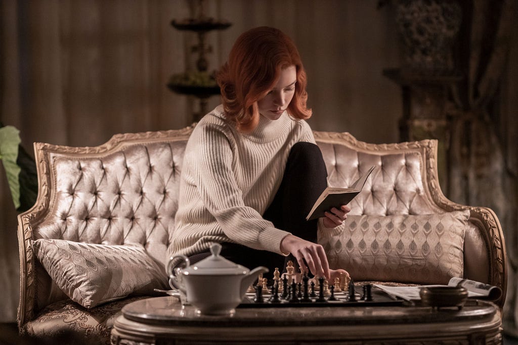 A woman sits on a couch, playing with the chess set in front of her while reading a book.