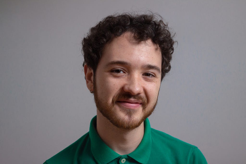 Portrait of a smiling 24-years-old white Spanish male, with short curly hair and short ginger beard. He is wearing a green polo shirt and he is standing in front of a mid gray wall. Only head and shoulders appear.