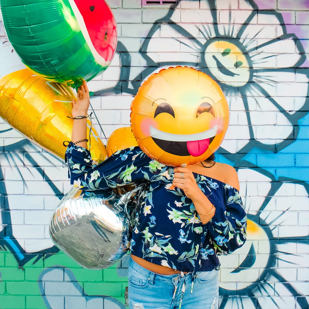 Person holding various balloons with a smiling, laughing emoji balloon covering their face