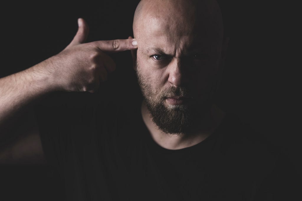 A man with a bald head and his hand making the shape of a pointing finger at his temple.