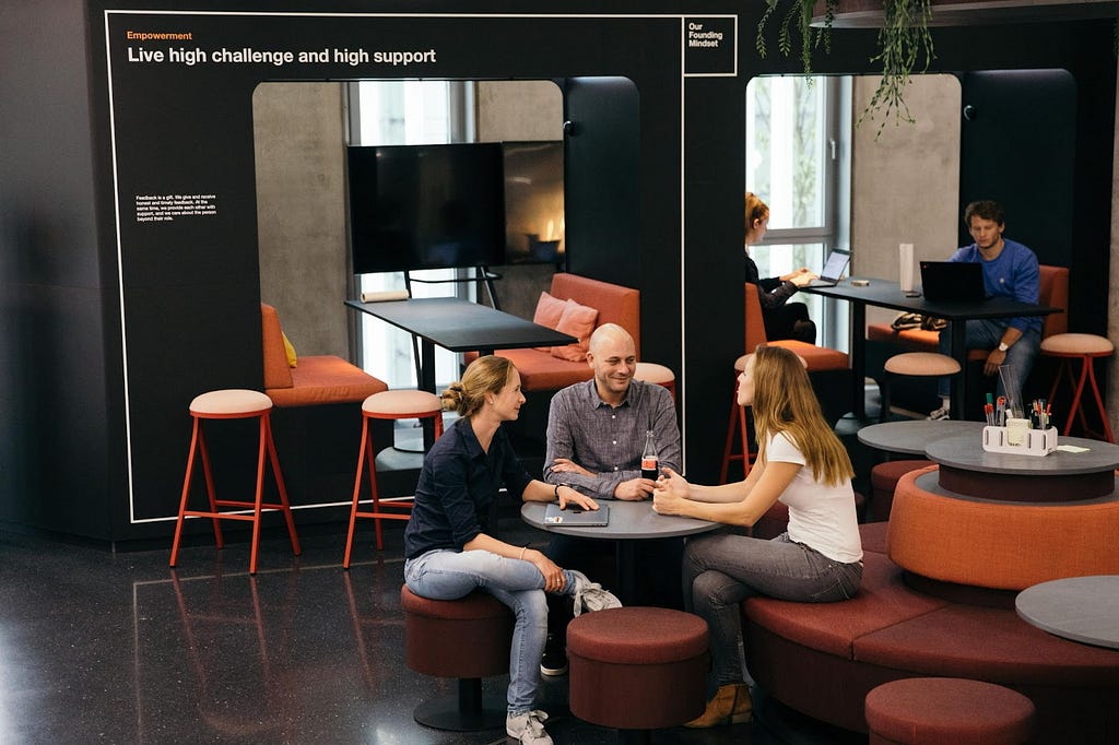 Members of the Zalando Product Design community sitting around a table at Zalando’s office cafeteria, talking and laughing. Behind them there’s a sign on one of the walls that reads “live high challenge and high support”.