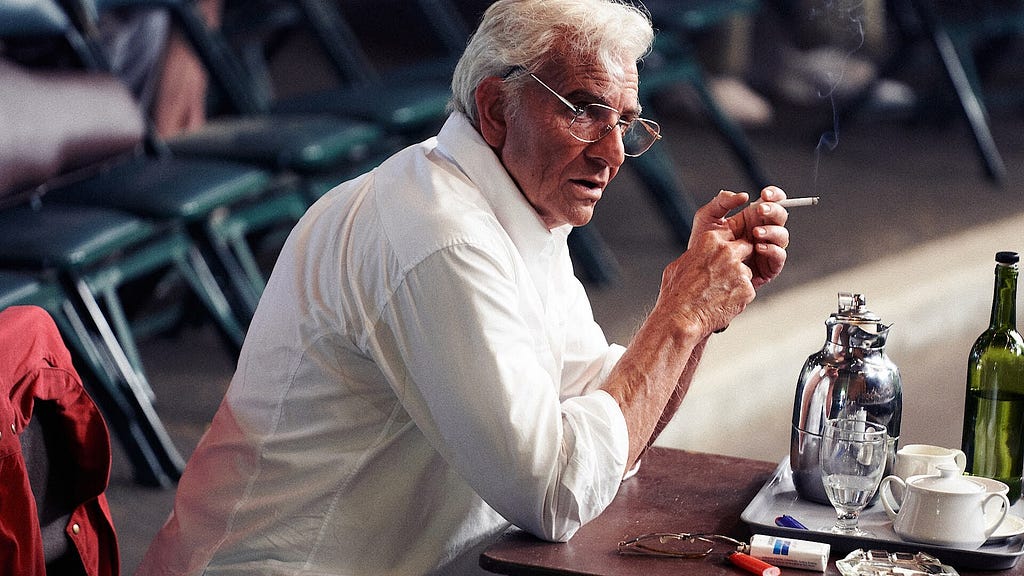 A man in glasses looks on intensely while smoking a cigarette in front of refreshments. (Bradley Cooper playing Leonard Bernstein)