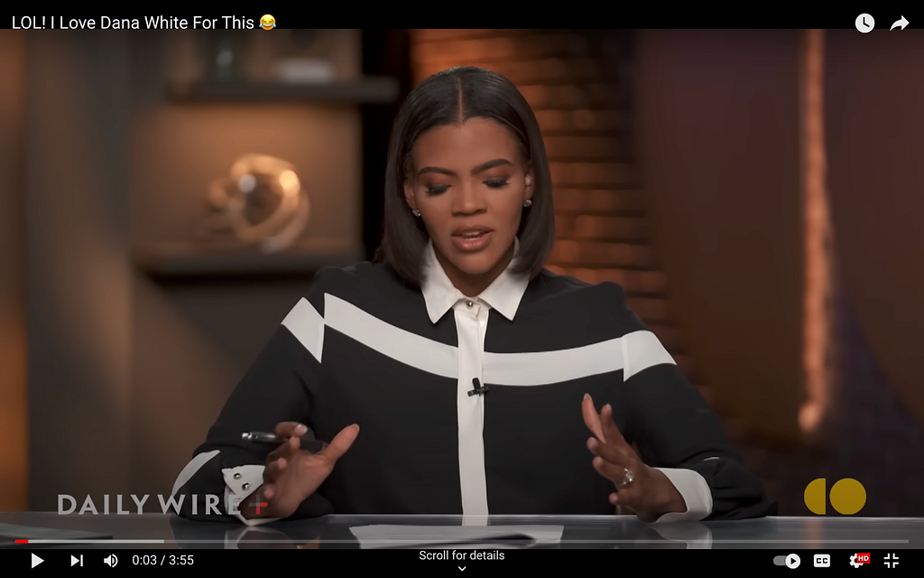 A screenshot from Candace Owens’s show on The Daily Wire. She’s in a somewhat flowy, long-sleeved blouse that’s black with a giant white cross across the front of it. The shirt collar is also white as well as pointed.