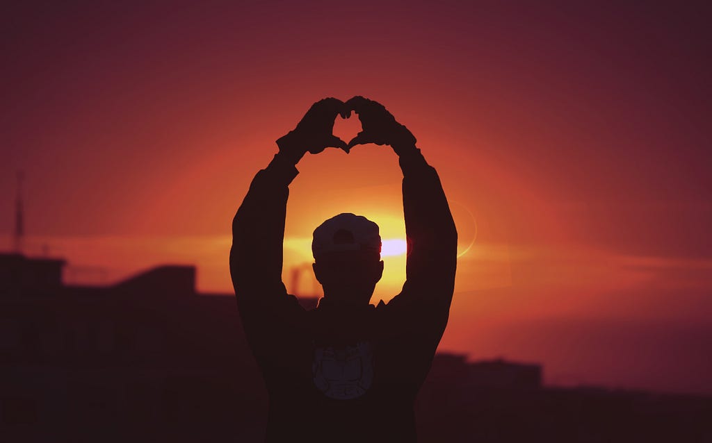 A person pictured at sunset. They are holding their hands above their head, using them to make a heart shape.