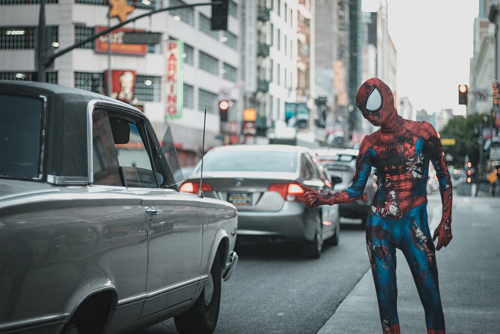 A beat-up Spiderman standing on the side of the road with his thumb up, looking to hitchhike a ride with a grey car.