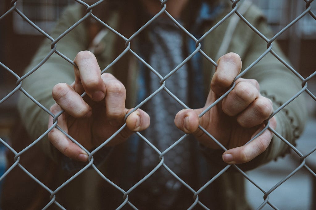 a person’s torso is shown standing behind a chain-link fence with their fingers gripping the metal. melancholy colour scheme.