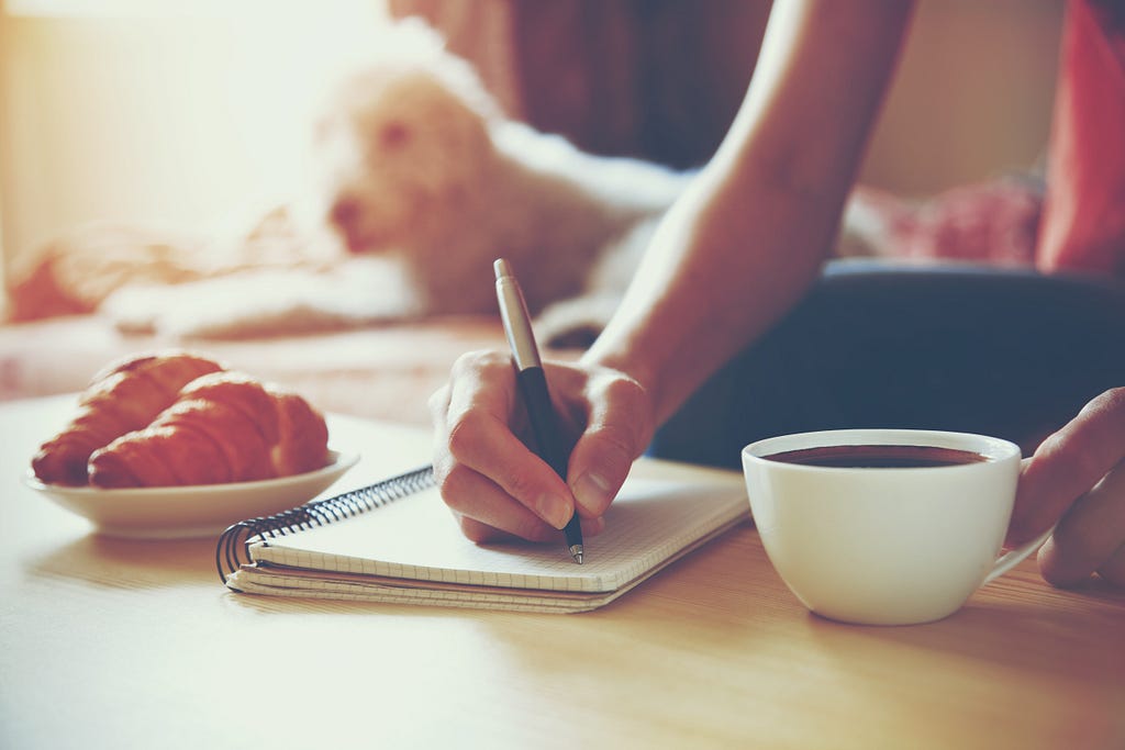 A woman writing on notebook with morning coffee and croissant.