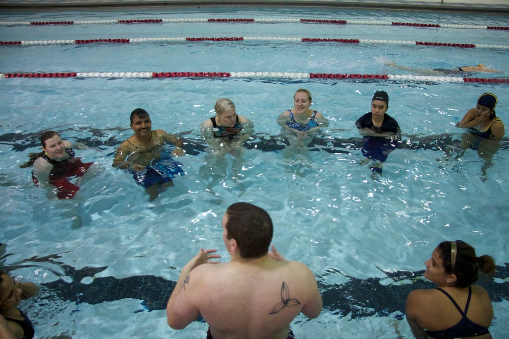 A group of 8 students gather around their swimming instructor during adult swimming lessons.