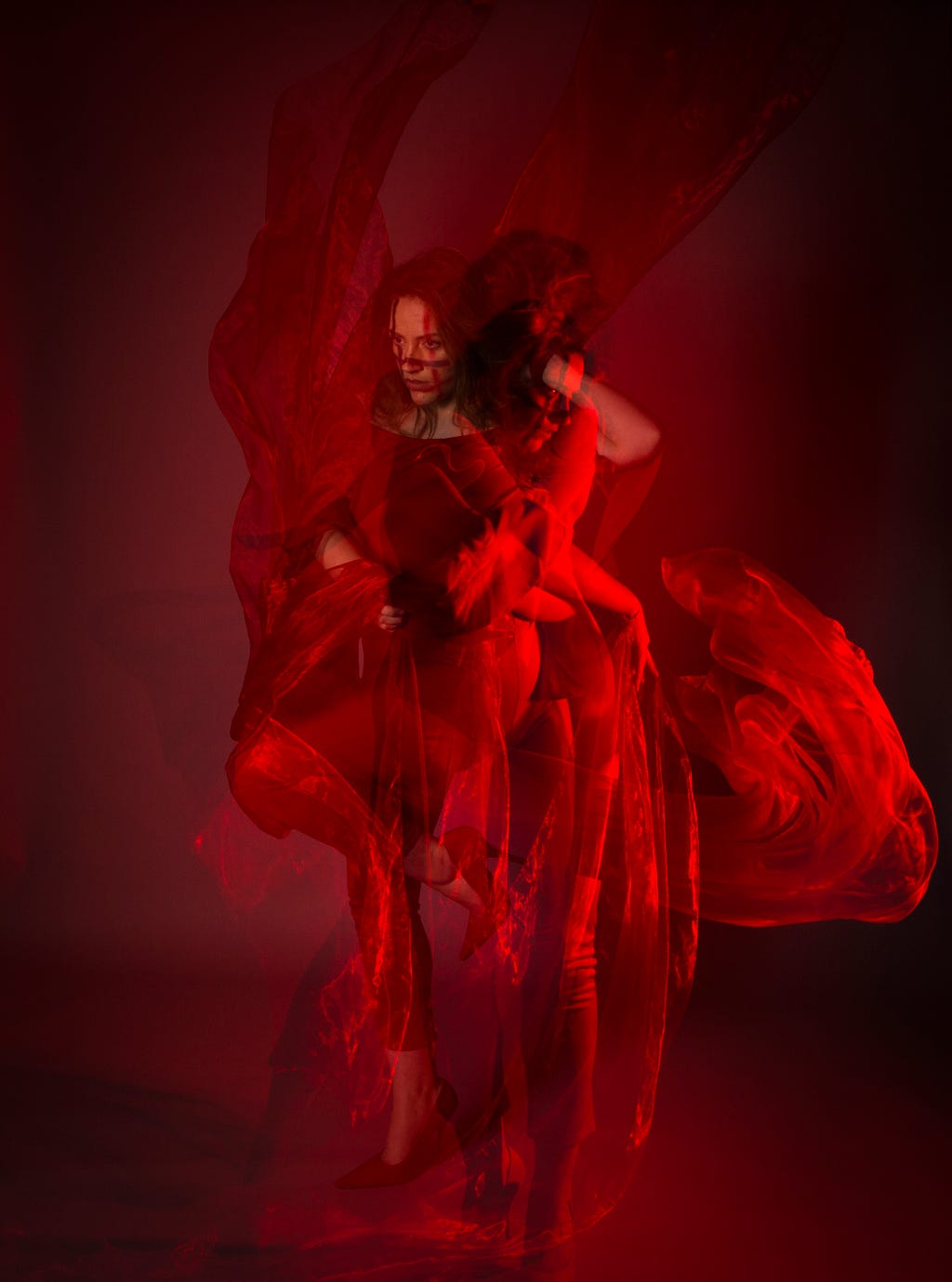 Multi-exposure photo of a brunette woman earing a red dress and scarves, against a dark red background. The fabric around her flows, and her movement is captured in the photo, with multiple angles of her in the image.