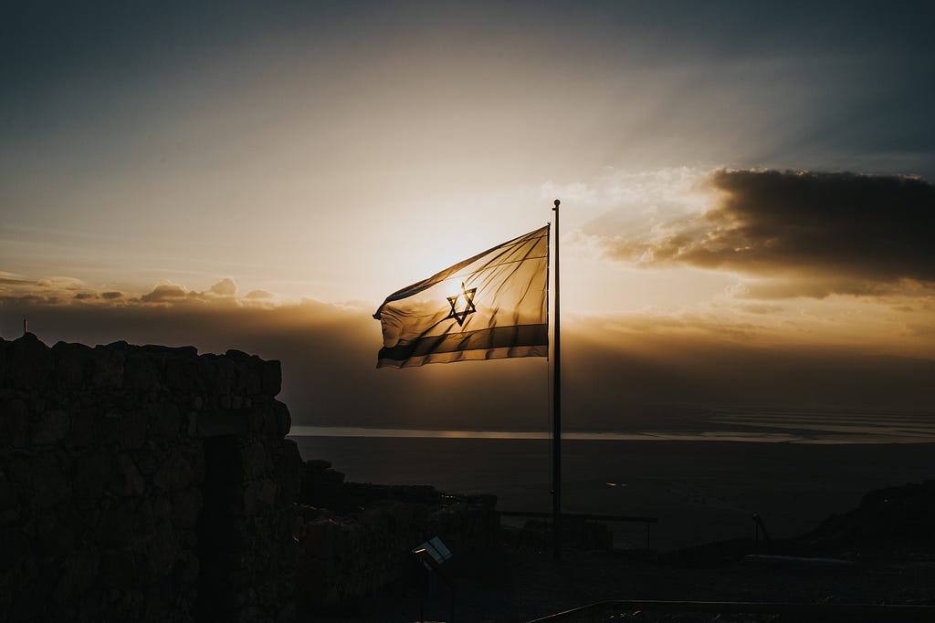 An Israeli flag waves in the wind against a dark sunset.